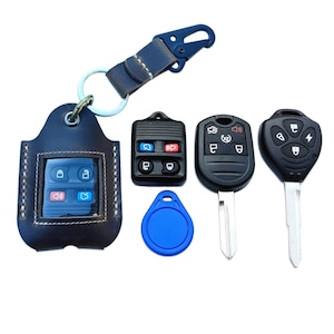 May include: A collection of car keys and key fobs. Includes a dark blue leather key holder with a clear window, several black key fobs, a blue circular tag, and a leather strap with a metal clasp.