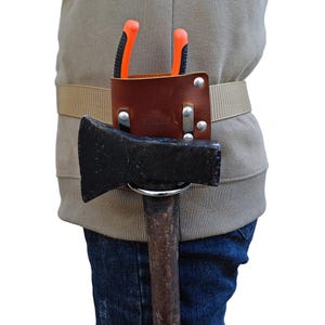 May include: A brown leather tool belt with an axe and pliers. The belt is secured with a beige strap. The axe has a black head and a wooden handle. The pliers have orange handles.
