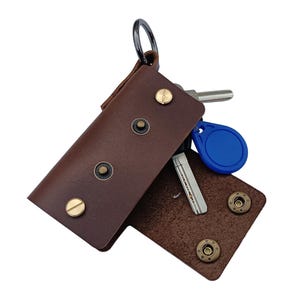 Könnte beinhalten: Ein brauner Leder-Schlüsselanhänger mit einem Metallring und mehreren Messing-Akzenten. Der Schlüsselanhänger ist geöffnet und zeigt einen blauen Schlüsselanhänger und einen silbernen Schlüssel im Inneren.