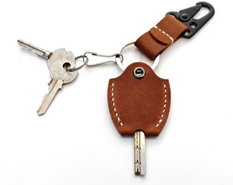 May include: A brown leather key organizer with white stitching and a silver metal snap button. The organizer holds a single key and is attached to a silver split ring with two additional keys and a black metal carabiner clip on a leather strap.