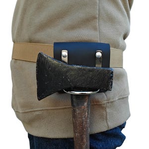 May include: A black leather axe sheath attached to a tan belt. The axe head is visible, with a wooden handle. The sheath is secured with silver-colored rivets. The person is wearing a tan hoodie and blue jeans.