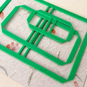 May include: Green plastic picture frame templates in various sizes, including 4x6, 3x3, and 1.5x1.5 inches, are stacked on a textured paper background with floral accents. The templates are designed for crafting and framing.