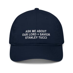 May include: A navy blue baseball cap with white embroidered text that reads "Ask me about our Lord + Savior Stanley Tucci".