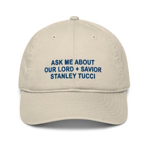 Peut inclure: Casquette de baseball beige avec l'inscription en bleu foncé "ASK ME ABOUT OUR LORD + SAVIOR STANLEY TUCCI". La casquette a une visière incurvée et une couronne arrondie.