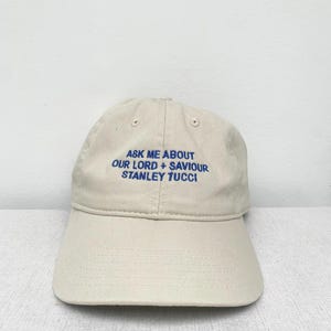 May include: A beige baseball cap with blue embroidered text that reads "Ask me about our lord + saviour Stanley Tucci".