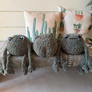 May include: Three handmade crochet frog plushies in varying poses. The frogs are a muted green color with large, blue eyes. They are resting on a patterned gray fabric, with cactus-themed pillows in the background.