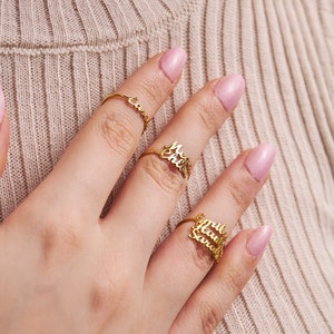May include: A hand wearing three gold rings with names on them. The names on the rings are "Cassie", "Melissa", and "Hailey Sarah".