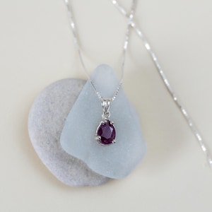 May include: A silver chain necklace with a pendant featuring a dark red gemstone. The pendant is set in a silver bezel with a delicate design.