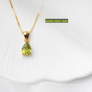 May include: A gold-colored necklace with a pear-shaped, light green gemstone pendant. The pendant is suspended from a delicate chain. The background is white.