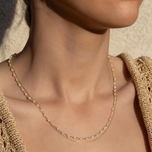 May include: A delicate gold chain necklace with small white beads. The necklace is layered, with two chains of different lengths.