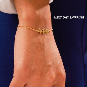 May include: A gold chain bracelet with a 444 charm. The bracelet is on a person's wrist.
