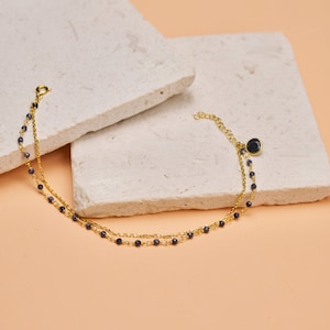 May include: A gold chain bracelet with a blue gemstone charm. The bracelet has two strands of chain with small blue gemstones spaced along the chain.