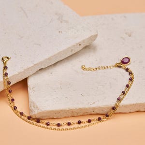May include: A gold chain bracelet with a single larger red gemstone charm and smaller purple gemstones along the chain.