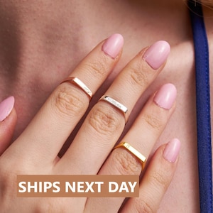 May include: Three gold rings with personalized engravings. The rings are stacked on a finger with pink nail polish. The top ring says "Olivia", the middle ring says "S", and the bottom ring says "Love".