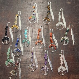 May include: A collection of handcrafted metal bookmarks, each featuring a tree of life design with colorful gemstone beads. The bookmarks have a silver feather charm attached. The gemstones vary in color, including black, yellow, blue, green, orange, and purple.