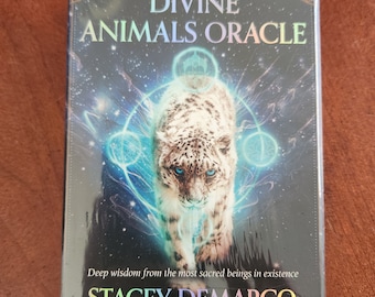 May include: A sealed oracle card deck titled "DIVINE ANIMALS ORACLE" featuring a snow leopard with blue eyes. The box has a dark background with a starry design and the author's name, STACEY DEMARCO, is printed at the bottom.