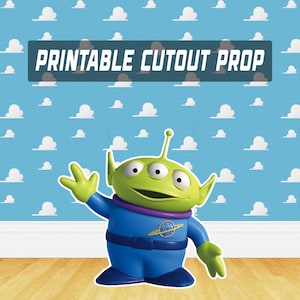 Peut inclure: Un accessoire de découpe imprimable représentant un extraterrestre vert à trois yeux, vêtu d'un uniforme bleu avec un col violet. Le fond est un ciel bleu avec des motifs de nuages blancs. Le texte "PRINTABLE CUTOUT PROP" est affiché.