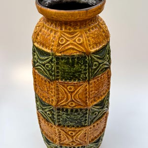May include: A tall, textured ceramic vase with a geometric pattern in shades of orange, green, and brown. The vase has a wide base and a narrower neck with a dark interior. The design features repeating squares and decorative motifs.