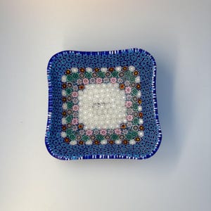 May include: Square glass dish with a blue floral pattern border and a white center. The dish has a slightly curved shape and is decorated with small, colorful floral designs. A decorative piece for the home.