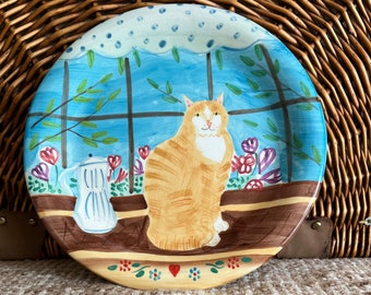 May include: A decorative ceramic plate featuring a whimsical design. The plate showcases a ginger cat sitting by a window with a blue sky background. The plate is adorned with floral and coffee pot motifs, adding a touch of charm.