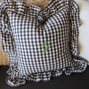 May include: A cushion with a black and white gingham design and a frilled edge. It has a green embroidered letter "J" in the centre. The cushion is on a black surface, with a fluffy, light-coloured background.