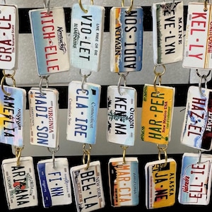 May include: A display of 12 pairs of earrings featuring miniature license plates from different states. Each earring is made of metal with a silver or gold hook. The license plates feature different names and state abbreviations. The earrings are displayed on a black stand.