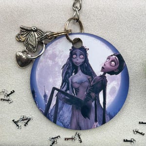 May include: A round, purple and blue key chain with a picture of Jack Skellington and Sally from the Nightmare Before Christmas movie. The keychain has a silver chain and a silver butterfly charm.