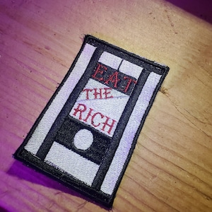 May include: A black and white embroidered patch with a guillotine design. The patch says "Eat the Rich" in red thread.