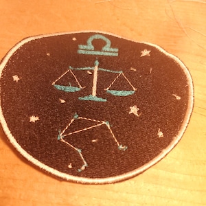 May include: A black and white embroidered patch with a teal blue zodiac symbol for Libra. The symbol is a set of scales with a constellation of stars in the background.
