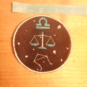 May include: A black and blue embroidered patch featuring the Libra zodiac symbol and constellation. The patch is circular and has a white outline.