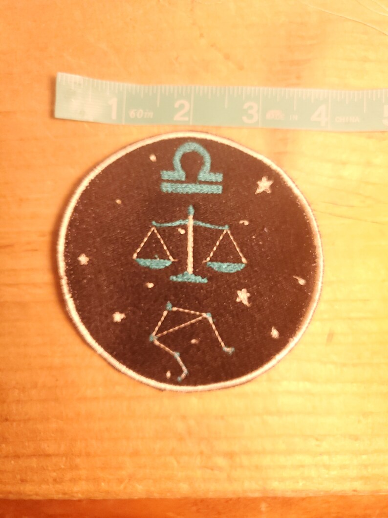May include: A black and blue embroidered patch featuring the Libra zodiac symbol and constellation. The patch is circular and has a white outline.