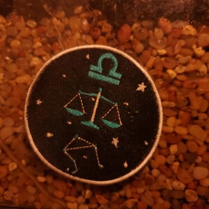 May include: A black and teal embroidered patch featuring the astrological symbol for Libra, a set of scales, and a constellation of stars.