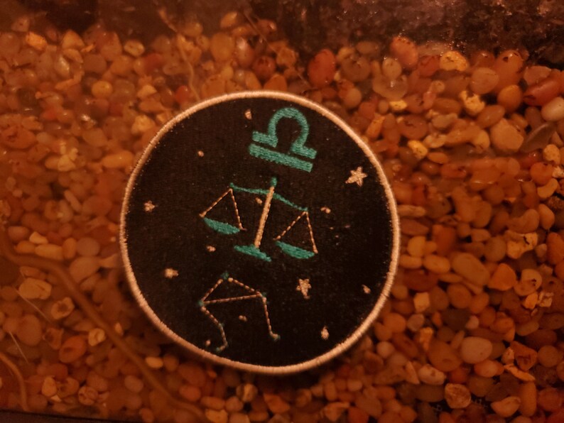 May include: A black and teal embroidered patch featuring the astrological symbol for Libra, a set of scales, and a constellation of stars.