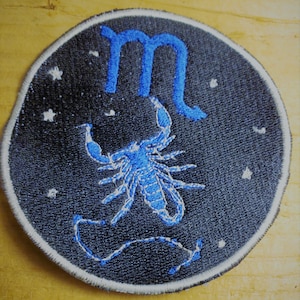 May include: A blue embroidered patch with a scorpion and the astrological symbol for Scorpio, against a black background with white stars.