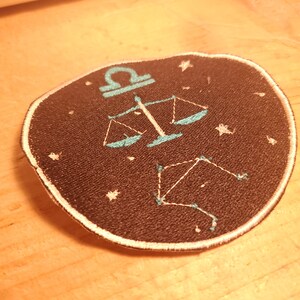 May include: A black embroidered patch with a white outline featuring the astrological symbol for Libra, a set of scales, and a constellation of stars in a teal blue colour.