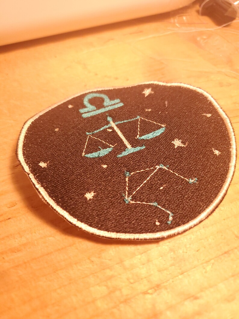 May include: A black embroidered patch with a white outline featuring the astrological symbol for Libra, a set of scales, and a constellation of stars in a teal blue colour.