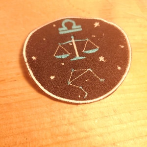 May include: A black and white embroidered patch featuring a blue Libra zodiac symbol and constellation on a black background.