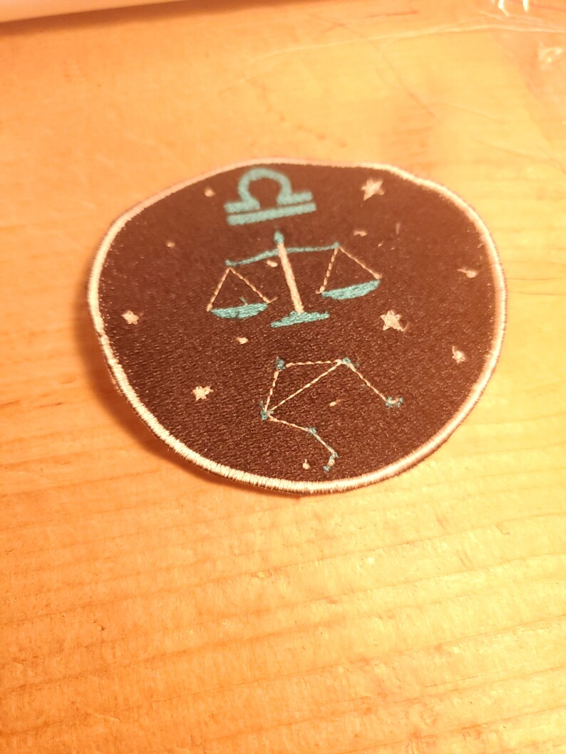 May include: A black and white embroidered patch featuring a blue Libra zodiac symbol and constellation on a black background.