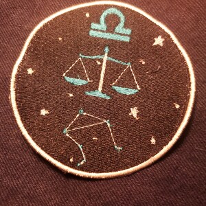May include: A round, black embroidered patch with a white outline. The patch features a teal blue Libra zodiac symbol with a constellation of stars.
