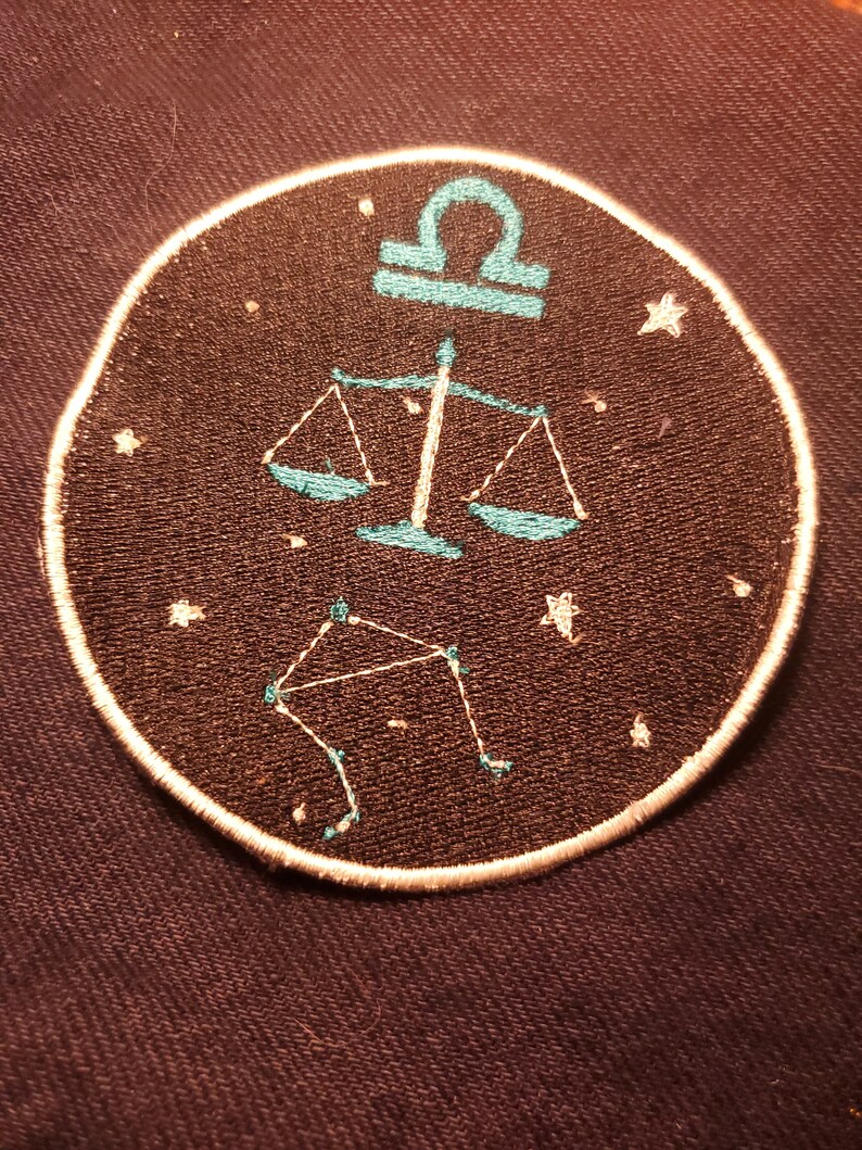 May include: A round, black embroidered patch with a white outline. The patch features a teal blue Libra zodiac symbol with a constellation of stars.
