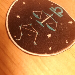 May include: A black circular patch with a white outline. The patch features a white outline of a constellation with a teal scale design in the center. The constellation is the Libra zodiac sign.