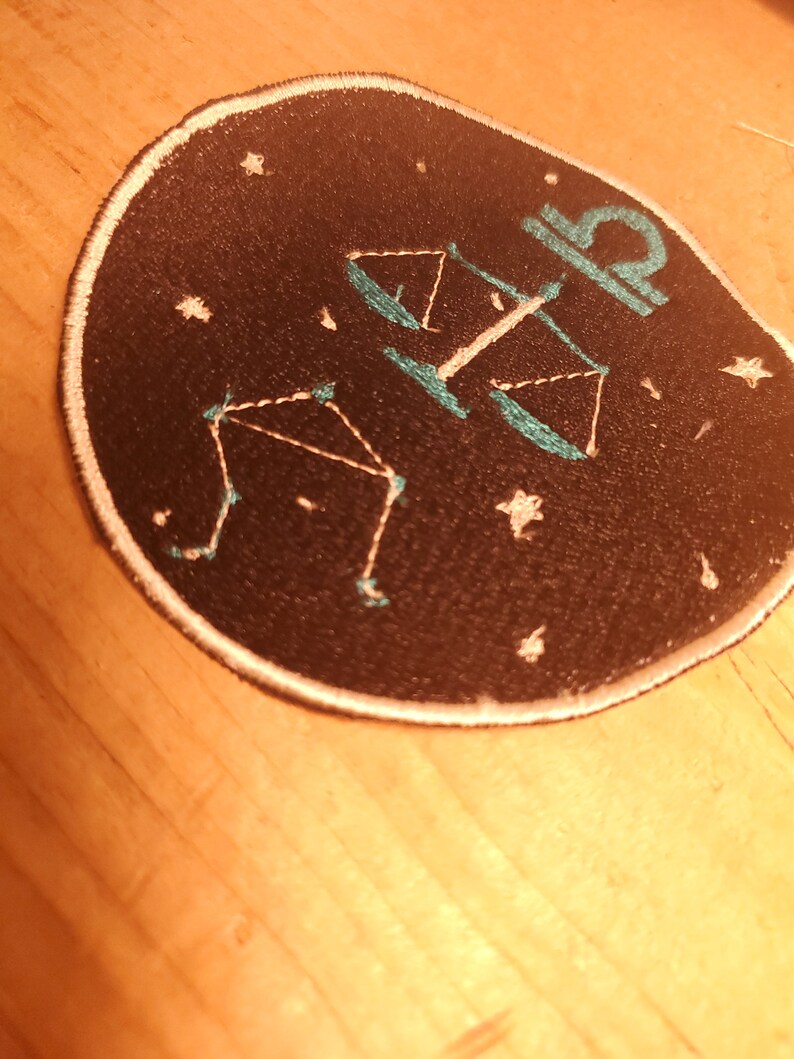 May include: A black circular patch with a white outline. The patch features a white outline of a constellation with a teal scale design in the center. The constellation is the Libra zodiac sign.