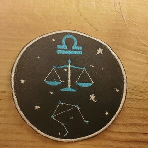 May include: A black and white embroidered patch featuring a blue scale design and the astrological symbol for Libra. The patch is surrounded by white stars on a black background.