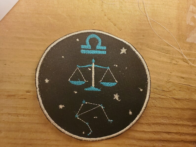 May include: A black and white embroidered patch featuring a blue scale design and the astrological symbol for Libra. The patch is surrounded by white stars on a black background.