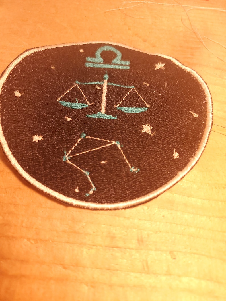 May include: A black and white embroidered patch featuring a teal blue zodiac symbol for Libra. The symbol is a set of scales with a constellation of stars in the background.