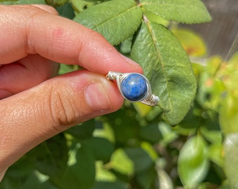 May include: A wire-wrapped ring featuring a single, round, deep blue lapis lazuli gemstone. The band is constructed from thin, silver-colored metal wire that coils around the stone to secure it in place, creating a textured, metallic setting.