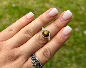 May include: A wire-wrapped ring featuring a round, polished tiger's eye gemstone in a golden-brown hue, paired with a second silver-toned ring that has a decorative, scalloped crown-like design with small circular cutouts along the band.