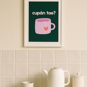 Puede incluir: Una impresión artística enmarcada con el texto "cupán tae?" encima de una ilustración de taza de té rosa con una bolsita de té en forma de corazón. Una tetera, taza, cuenco y bote blancos están sobre una encimera blanca.