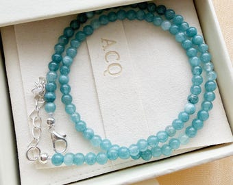 Puede incluir: Un collar hecho de pequeñas cuentas redondas y translúcidas de color azul verdoso dispuestas en un patrón circular. El collar cuenta con un cierre de langosta de metal plateado y una cadena extensora ajustable, dentro de una caja blanca con el texto ACQ.