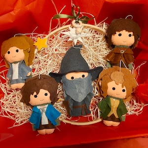 May include: A set of five felt character ornaments featuring a central wizard in a blue hat, surrounded by four smaller figures with brown, blonde, and dark hair, all arranged around a wooden embroidery hoop with a white star and green ribbon.
